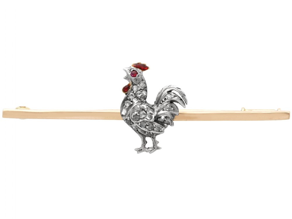An antique 14ct yellow gold bar brooch adorned with a silver-set diamond cockerel motif featuring a ruby eye and red enamelled details.