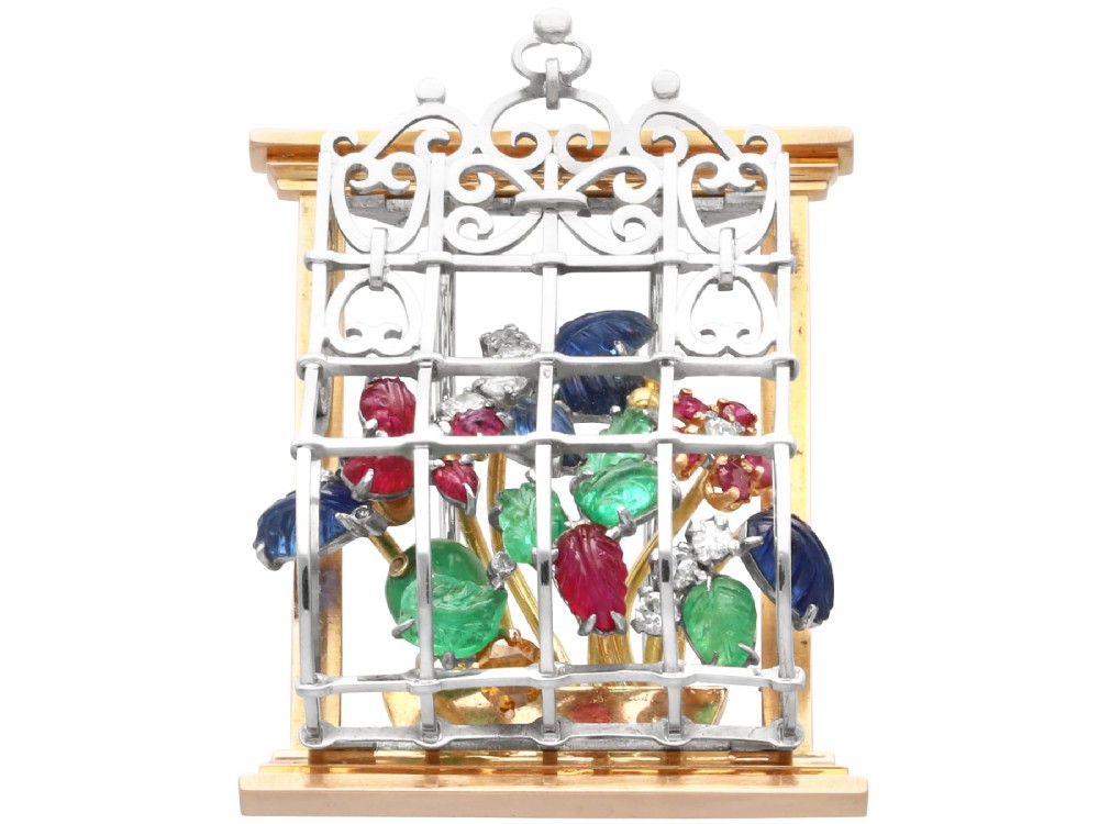 An 18ct gold window-framed brooch featuring carved emerald, ruby, and sapphire floral elements behind a silver-coloured metal grate.