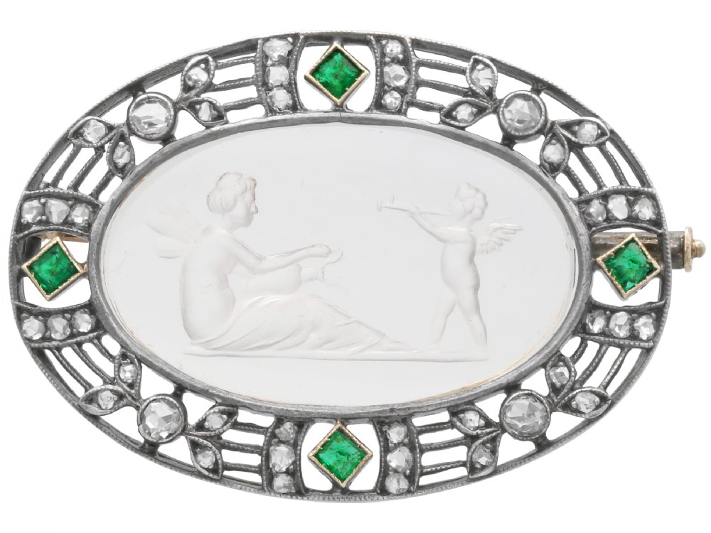 An oval Victorian brooch featuring a reverse-carved rock crystal intaglio depicting Cupid and Psyche, bordered by rose-cut diamonds and square-cut emeralds set in gold and silver.