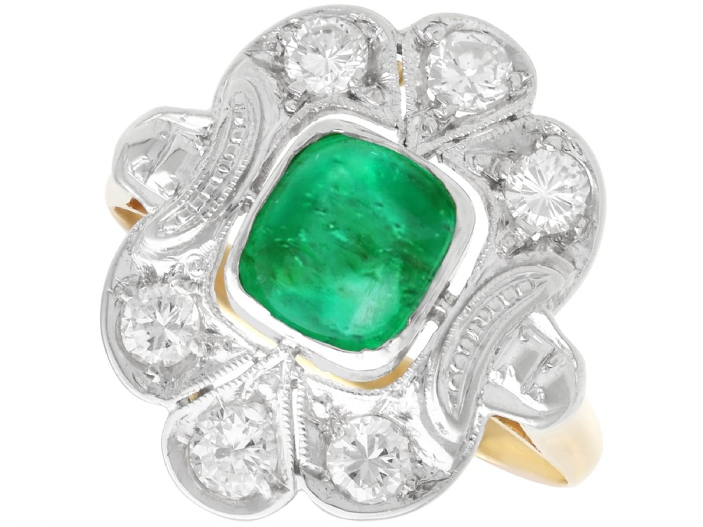 An Art Deco dress ring featuring a central cushion-cut green emerald surrounded by a geometric pierced diamond halo set in white metal on an 18ct yellow gold band.