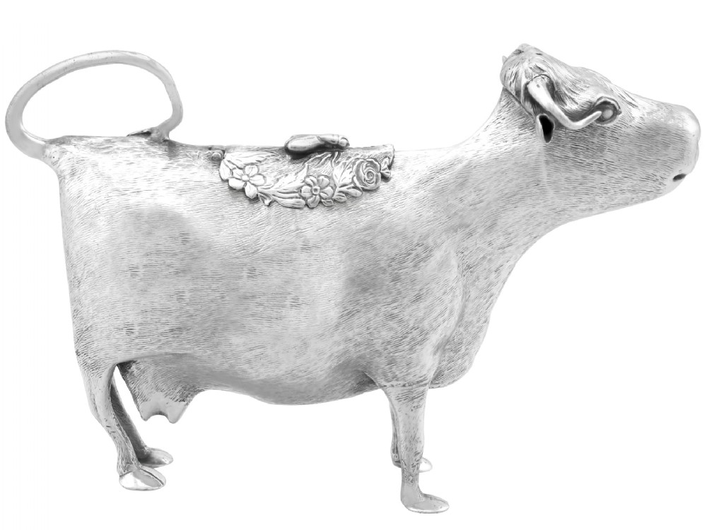 An antique sterling silver cow creamer featuring textured hide detailing, a looped tail handle, and a floral hinged lid on its back.
