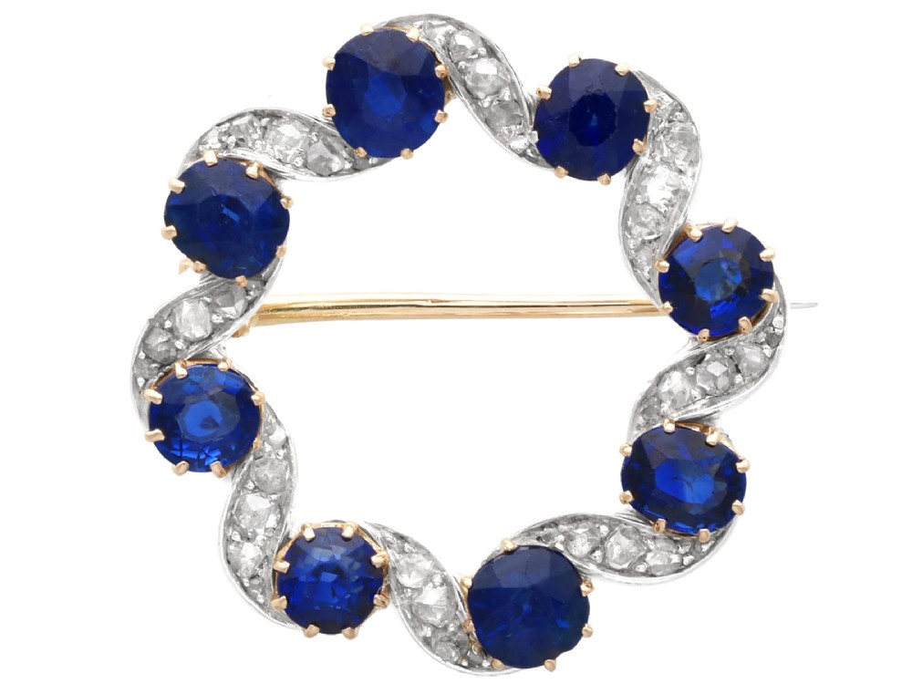 A circular late Victorian wreath brooch set with eight oval blue sapphires and diamond-set silver ribbon scroll motifs on a yellow gold frame.