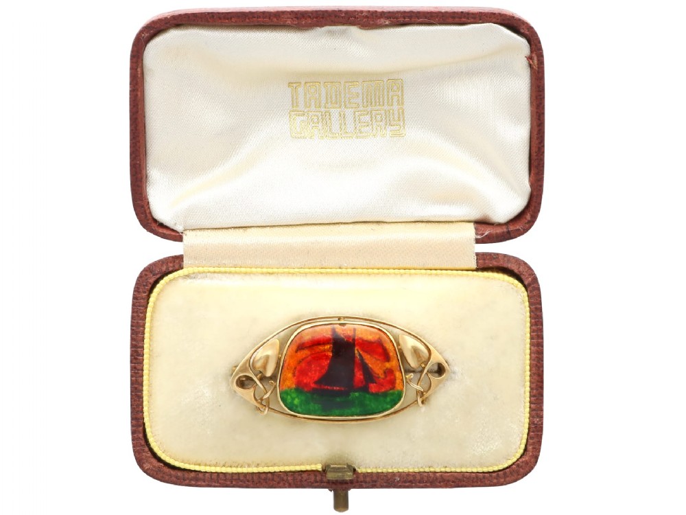 An Arts and Crafts 15ct yellow gold brooch featuring a central enamel panel depicting a sailboat against a sunset sky framed by stylized gold leaves.