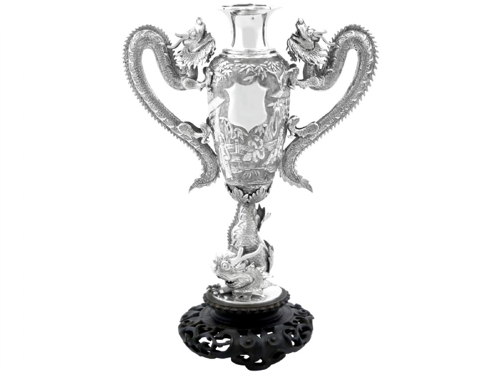 A silver trophy-style vase featuring sculpted dragon handles, relief carving, and a dark wooden base.