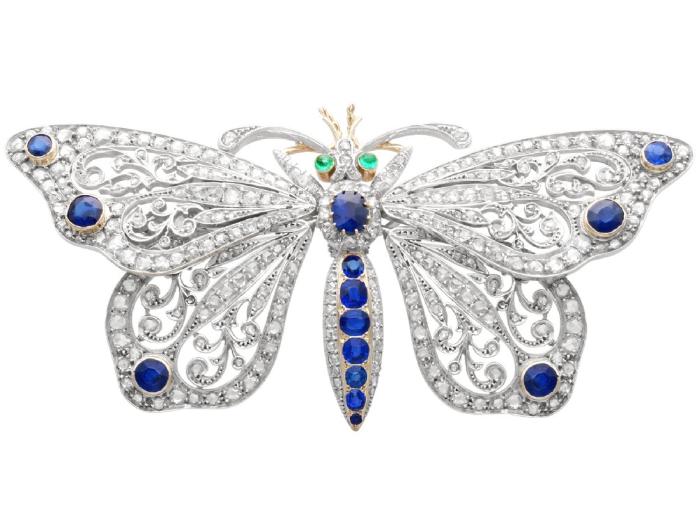 An antique butterfly brooch constructed from gold and silver, featuring openwork wings set with diamonds, sapphire details, and green emerald eyes.