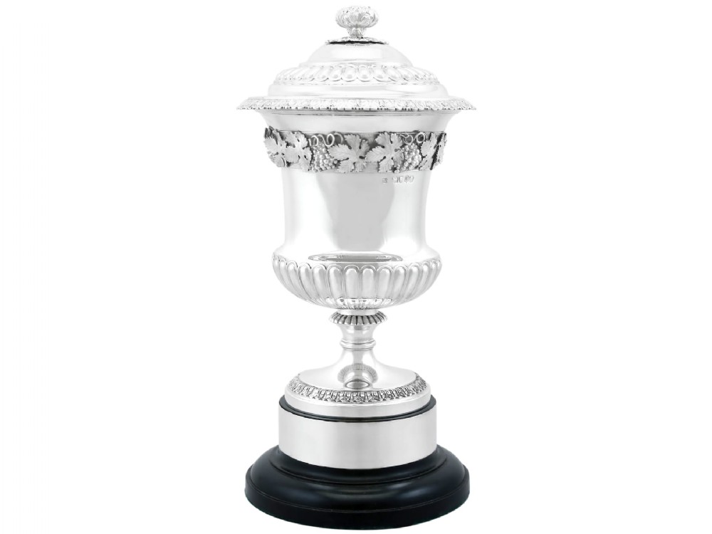 A Victorian sterling silver presentation cup and cover decorated with a grape leaf band, resting on a round ebonised wood plinth.
