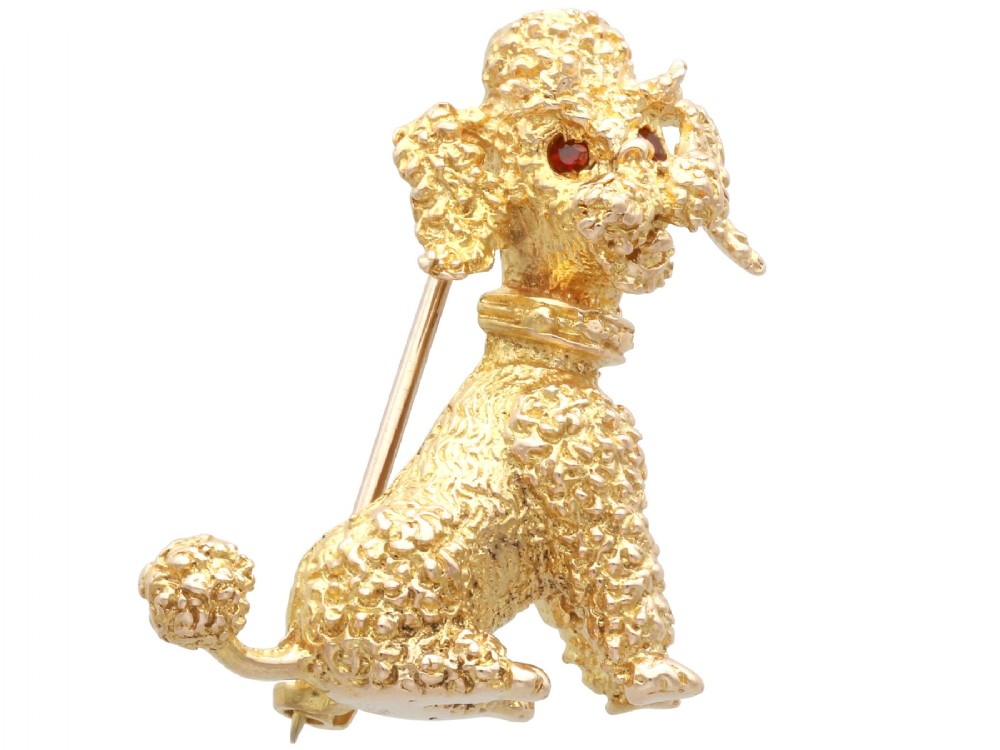 A 9ct yellow gold brooch sculpted as a seated poodle with textured fur and red garnet eyes holding a stick in its mouth.