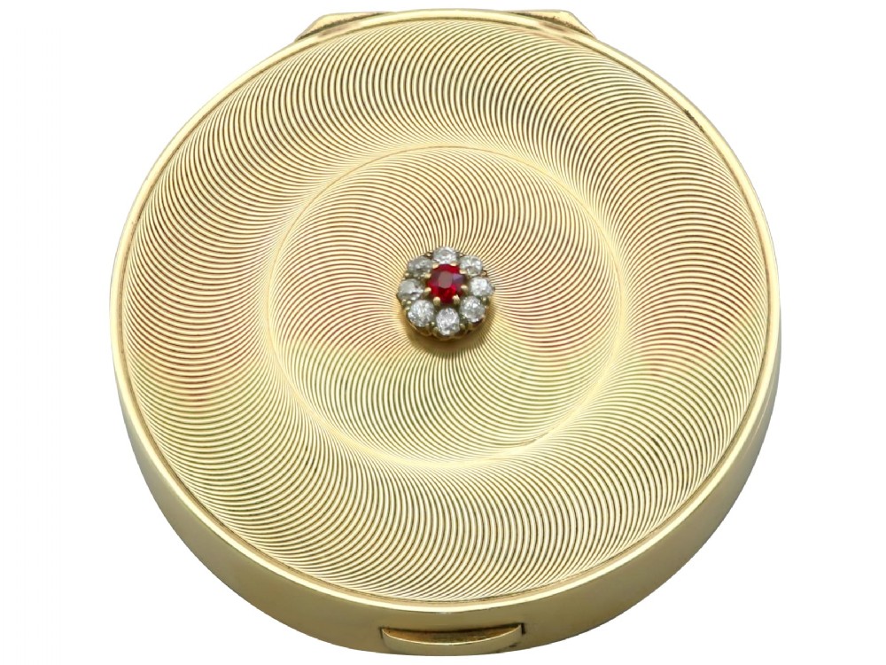 A circular 9ct yellow gold powder compact featuring an engine-turned swirling pattern and a central ruby and diamond cluster.