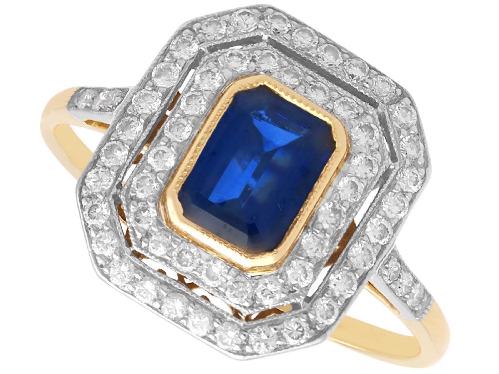 An 18ct yellow gold ring featuring a central emerald-cut blue sapphire framed by a geometric double halo of round diamonds with millegrain edging.