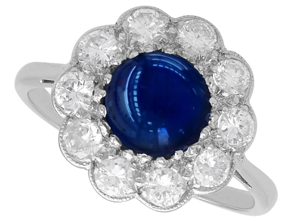 A platinum ring set with a central round cabochon blue sapphire surrounded by a halo of ten round diamonds.