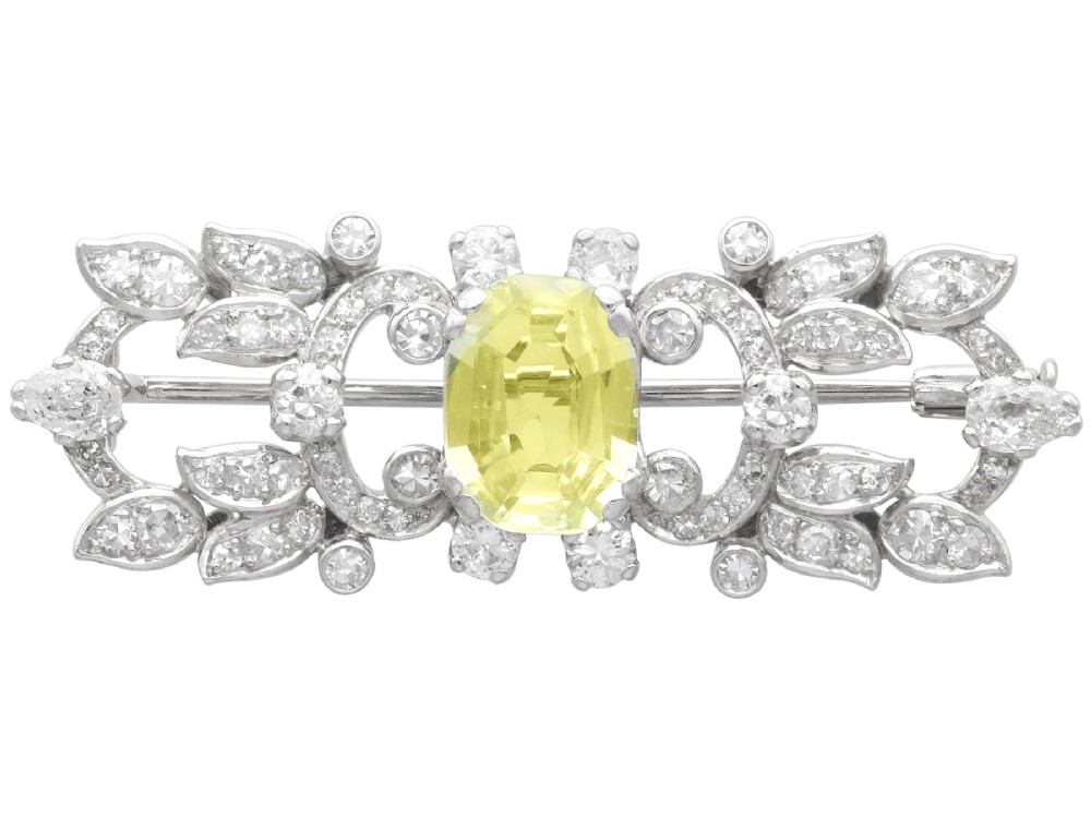 An Art Deco platinum brooch centered with an oval cushion-cut yellow sapphire enclosed by diamond-set scrollwork and foliate motifs.