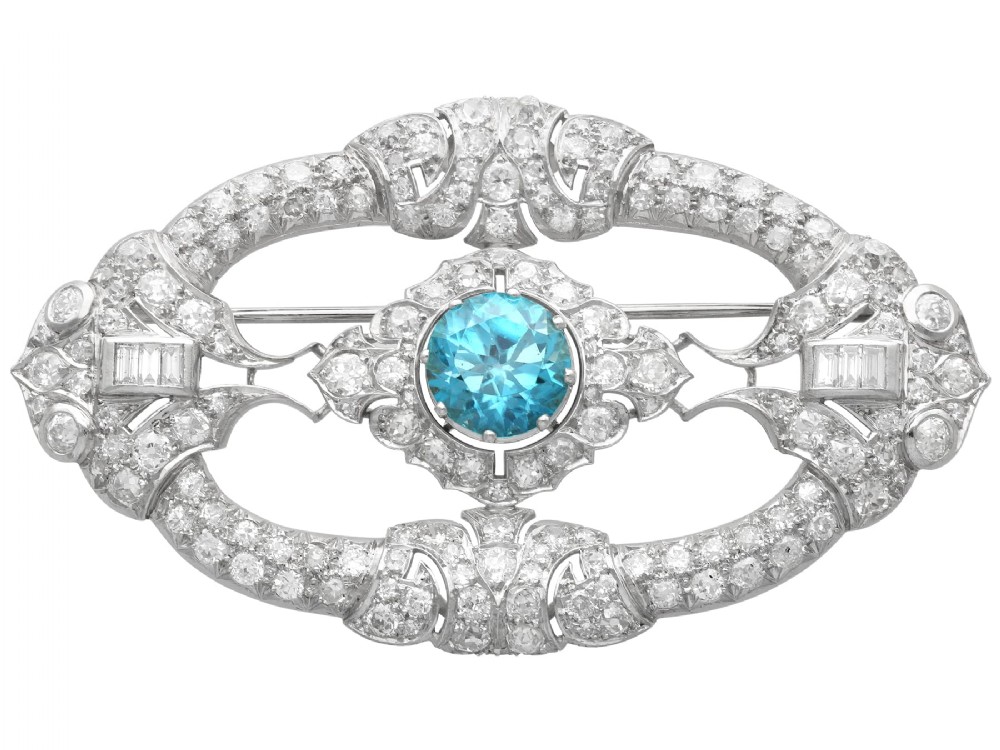 An oval Art Deco platinum brooch featuring an openwork geometric frame set with diamonds and a central round brilliant blue zircon.