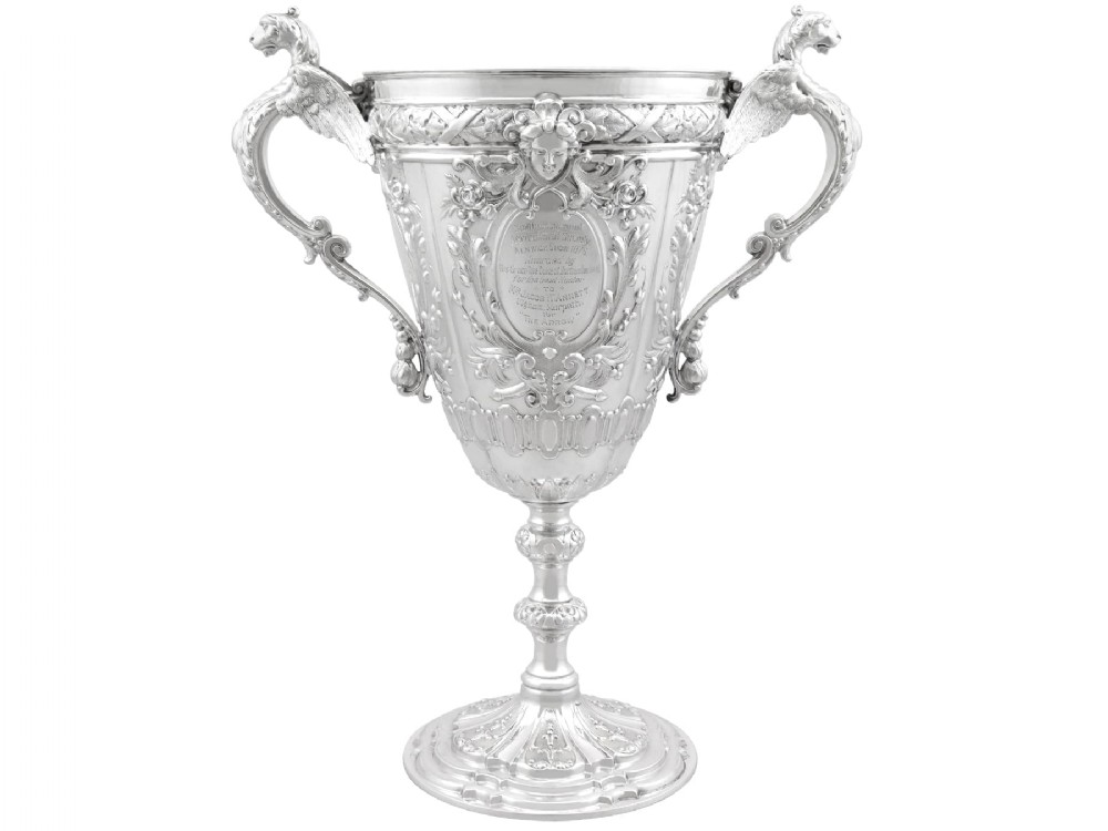An antique Victorian sterling silver presentation cup featuring ornate chased scrollwork, twin handles topped with winged figures, and a stepped pedestal base.