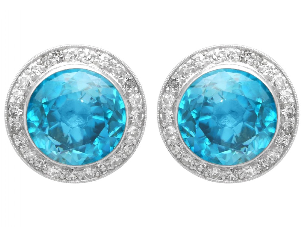 A pair of 14ct white gold stud earrings featuring round blue zircon gemstones encircled by diamond halos with millegrain borders.