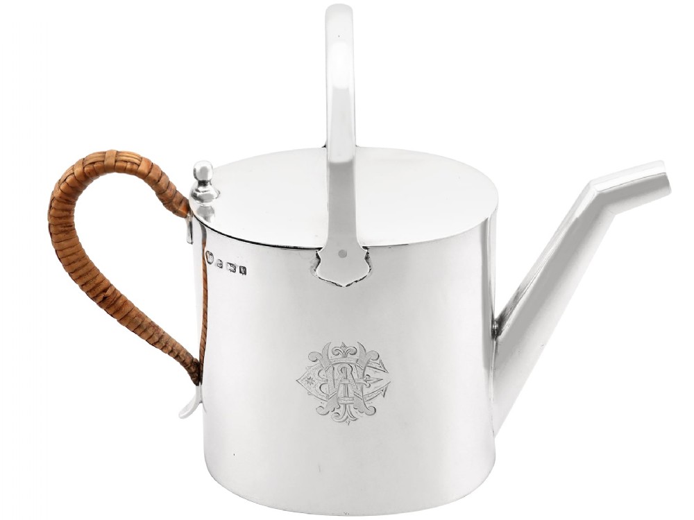 An Edwardian sterling silver watering can featuring a cylindrical body, straight spout, overhead loop handle, woven cane side handle, and an engraved central monogram.
