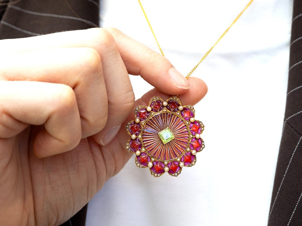 An Edwardian 15ct yellow gold circular pendant featuring radial red and pink enamel, set with a central square peridot and bordered by seed pearls. View V