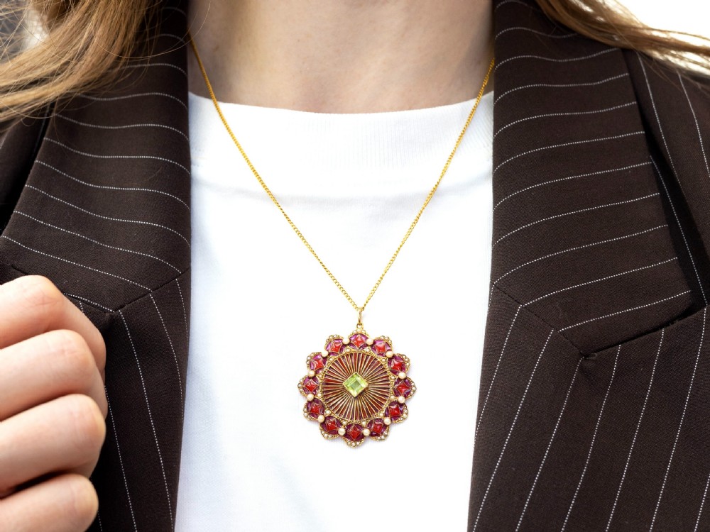 An Edwardian 15ct yellow gold circular pendant featuring radial red and pink enamel, set with a central square peridot and bordered by seed pearls. View VI