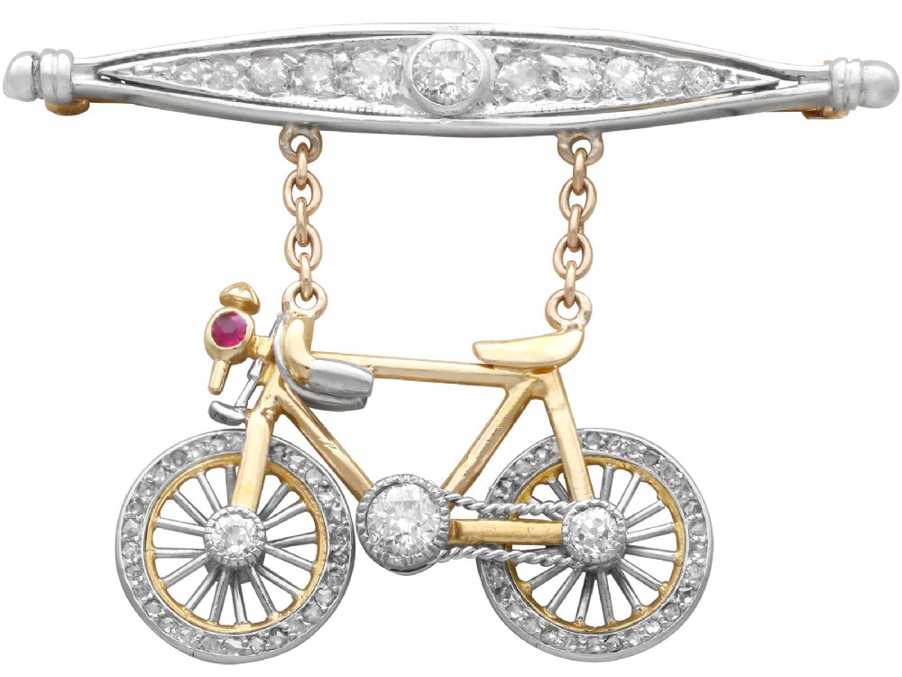 An antique Edwardian bar brooch featuring a suspended yellow gold bicycle with diamond-set wheels, set beneath a platinum-faced bar set with diamonds.