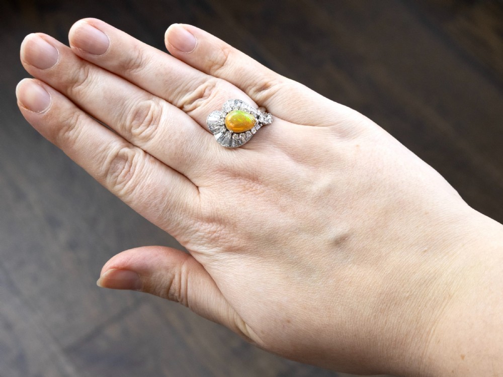 A platinum dress ring featuring a central pear-shaped orange opal surrounded by a halo of round and baguette-cut diamonds in an asymmetrical frame. View VII