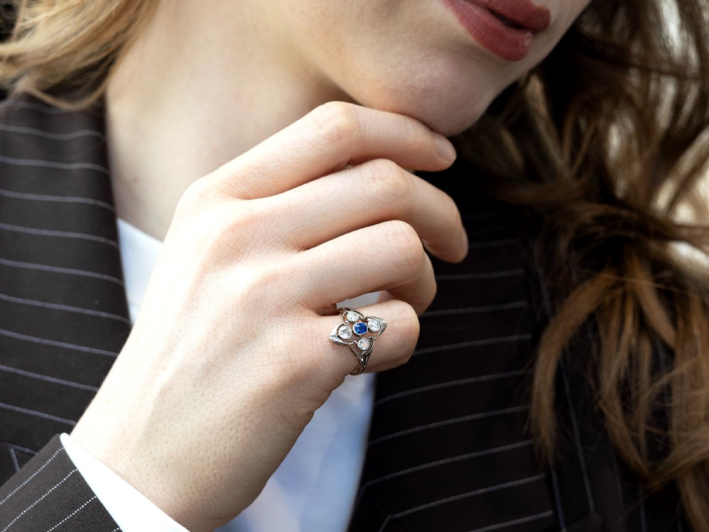 An Art Deco 14ct yellow gold ring featuring a central oval blue sapphire surrounded by four round diamonds set in a diamond-shaped openwork frame. View IV