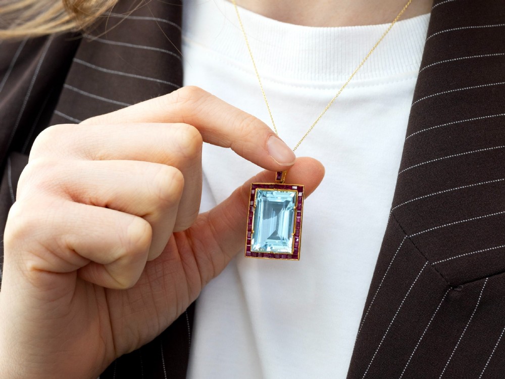 A large rectangular step-cut blue aquamarine set in yellow gold with a square-cut red ruby border and ruby-set bail. View IV