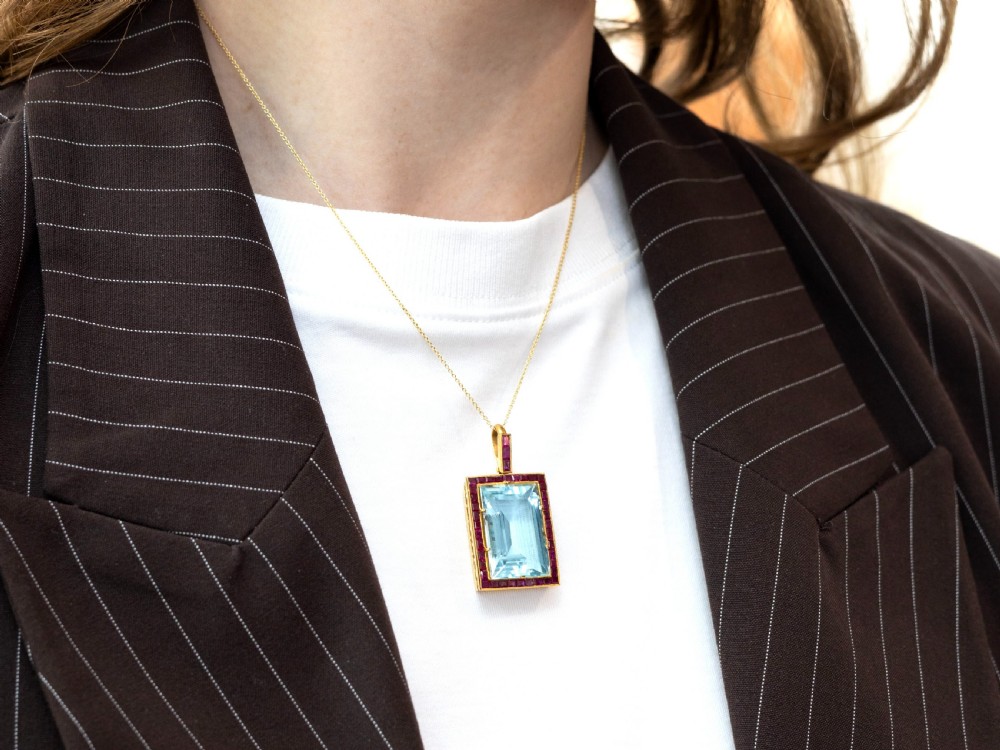 A large rectangular step-cut blue aquamarine set in yellow gold with a square-cut red ruby border and ruby-set bail. View V