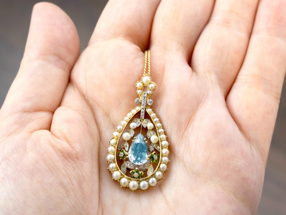 An openwork Edwardian 14ct yellow gold pendant set with a central pear-cut aquamarine surrounded by seed pearls, diamonds, and green demantoid garnets. View V