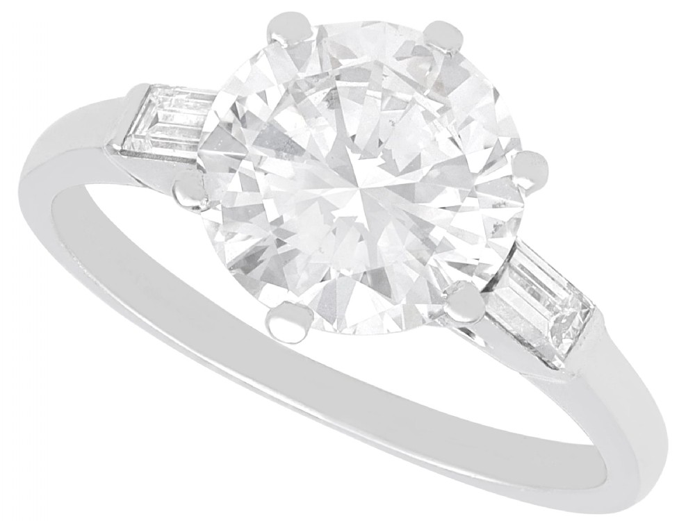 A platinum solitaire ring featuring a large round cut central diamond set with six claws, flanked by step-cut baguette diamonds on each shoulder.