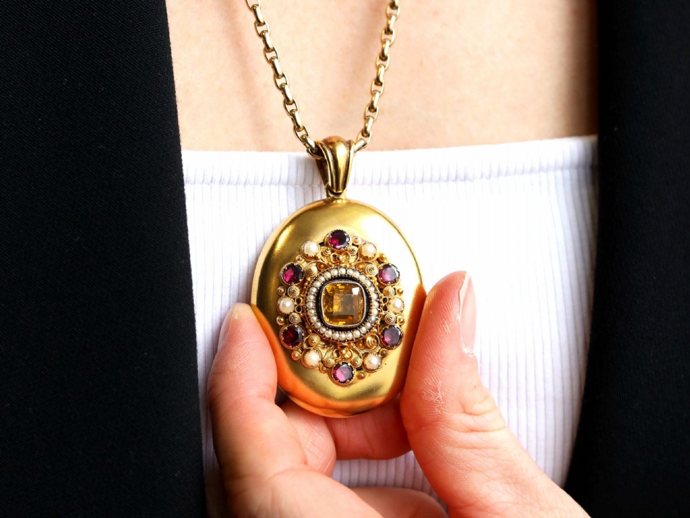 An oval 18ct yellow gold Victorian locket featuring a central yellow citrine surrounded by seed pearls, garnets, and detailed gold wirework. View V