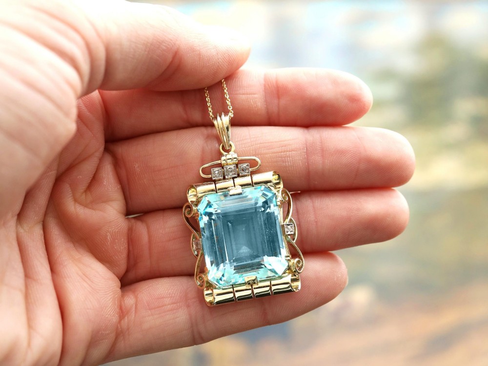 A large emerald-cut aquamarine gemstone mounted in a yellow gold scrollwork frame accented with small diamonds and suspended from a chain. View III