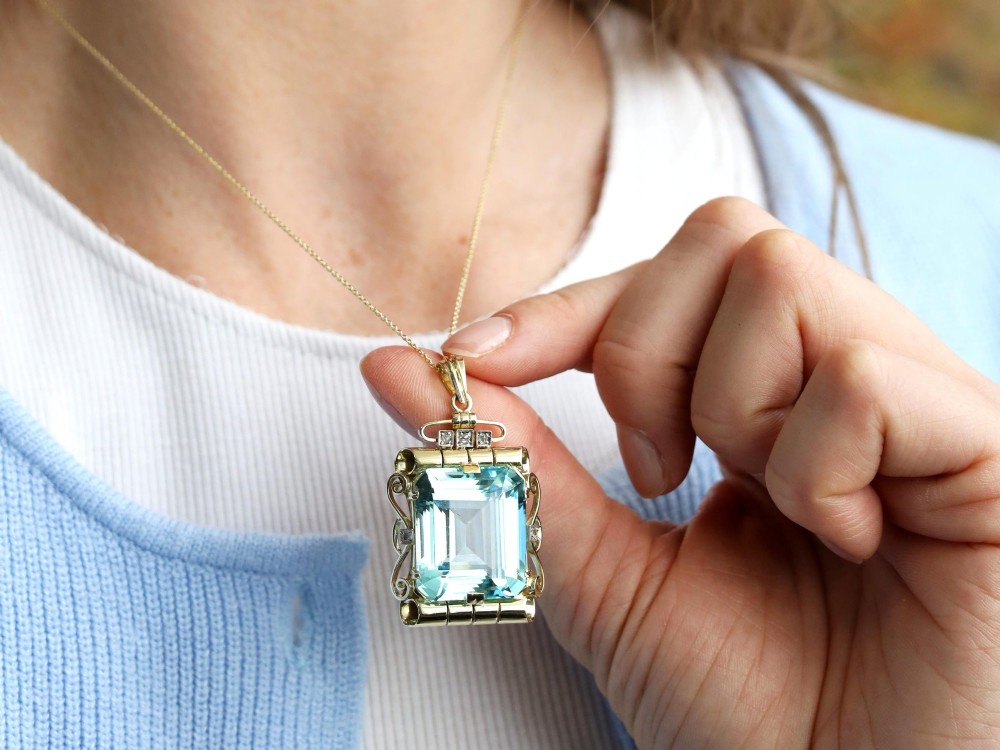 A large emerald-cut aquamarine gemstone mounted in a yellow gold scrollwork frame accented with small diamonds and suspended from a chain. View IV