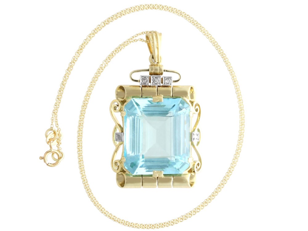 A large emerald-cut aquamarine gemstone mounted in a yellow gold scrollwork frame accented with small diamonds and suspended from a chain. View VI