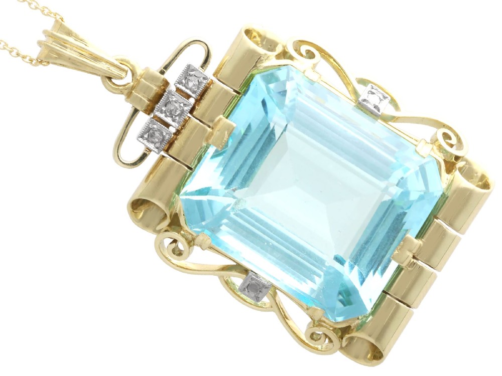 A large emerald-cut aquamarine gemstone mounted in a yellow gold scrollwork frame accented with small diamonds and suspended from a chain. View VII