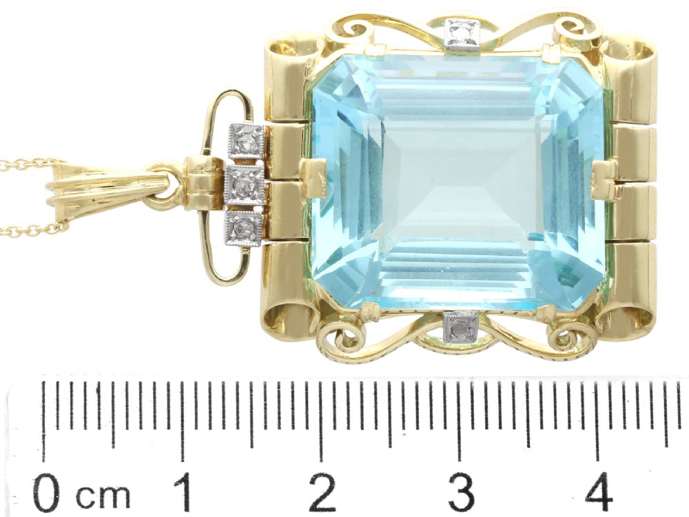 A large emerald-cut aquamarine gemstone mounted in a yellow gold scrollwork frame accented with small diamonds and suspended from a chain. View X