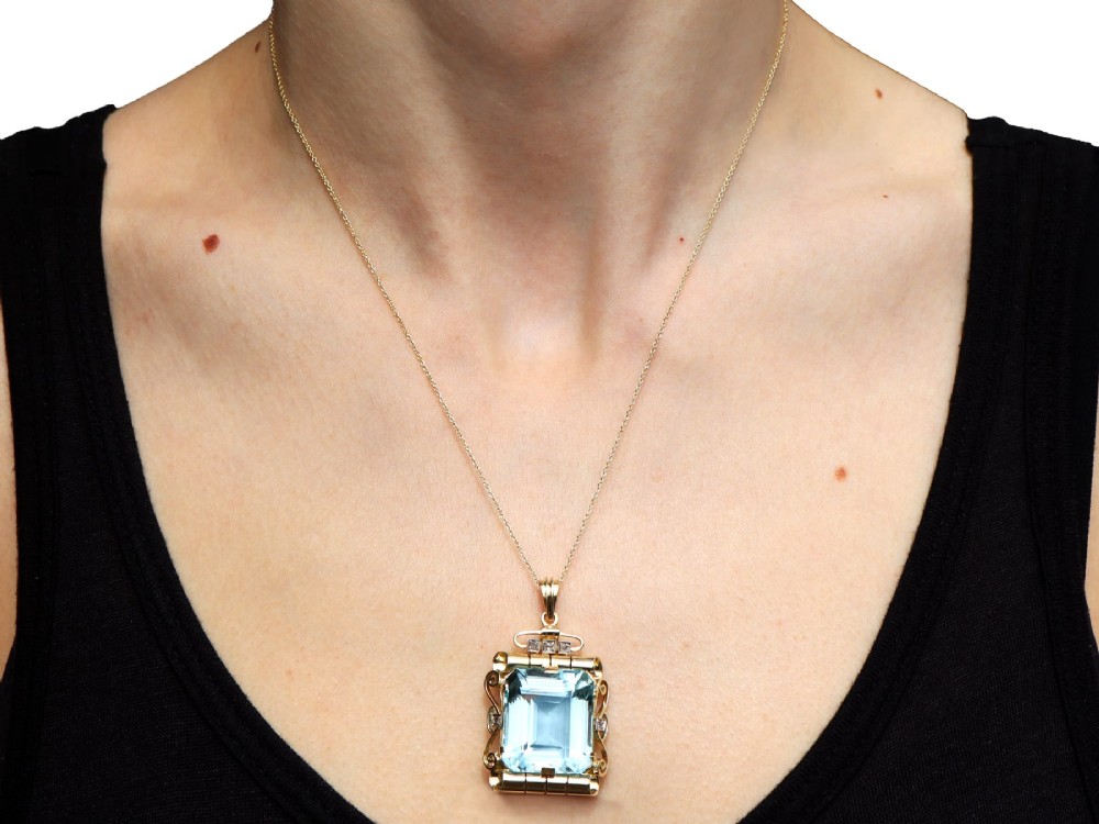 A large emerald-cut aquamarine gemstone mounted in a yellow gold scrollwork frame accented with small diamonds and suspended from a chain. View XII