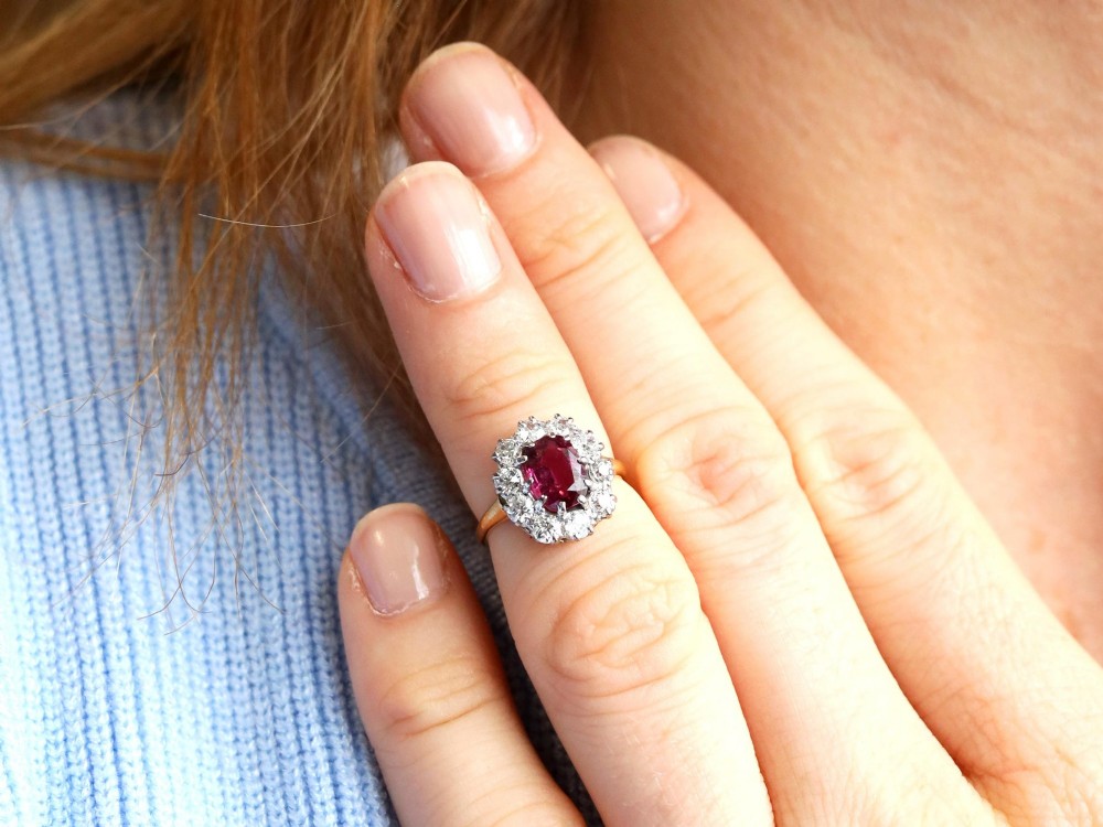 An antique 18ct yellow gold ring featuring a central oval red ruby set within a cluster halo of round white diamonds. View V