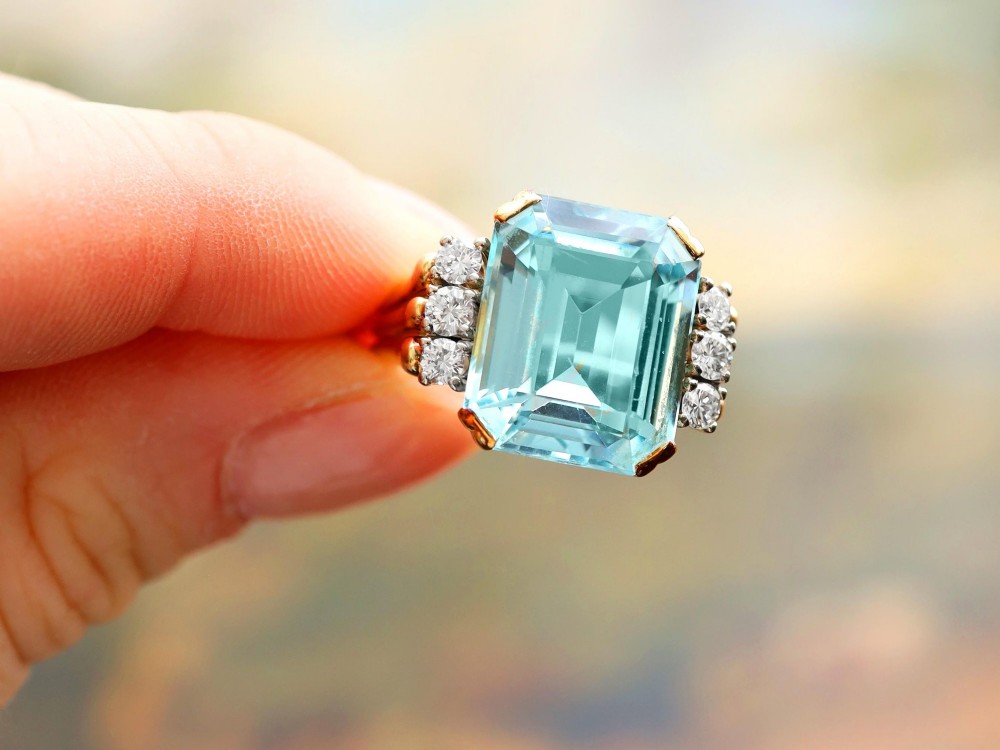 A 14ct yellow gold dress ring featuring a large emerald-cut light blue aquamarine central gemstone flanked by six round diamonds. View V