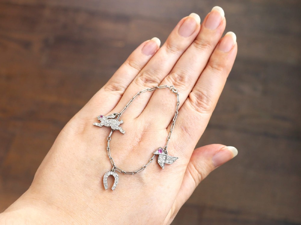 An antique platinum link chain charm bracelet featuring diamond-set charms including a running hare, a horseshoe, and a bird with ruby accents. View V