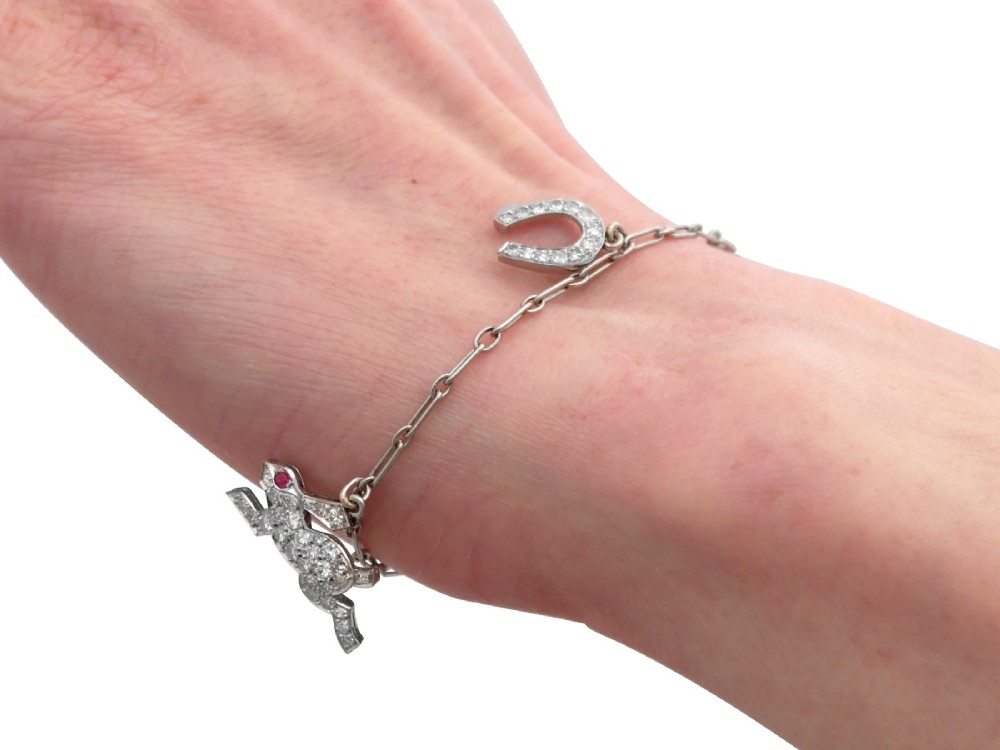 An antique platinum link chain charm bracelet featuring diamond-set charms including a running hare, a horseshoe, and a bird with ruby accents. View XI