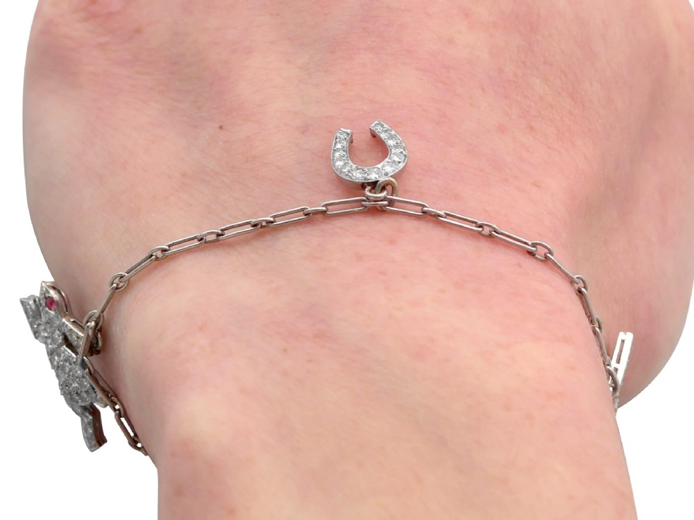 An antique platinum link chain charm bracelet featuring diamond-set charms including a running hare, a horseshoe, and a bird with ruby accents. View XII