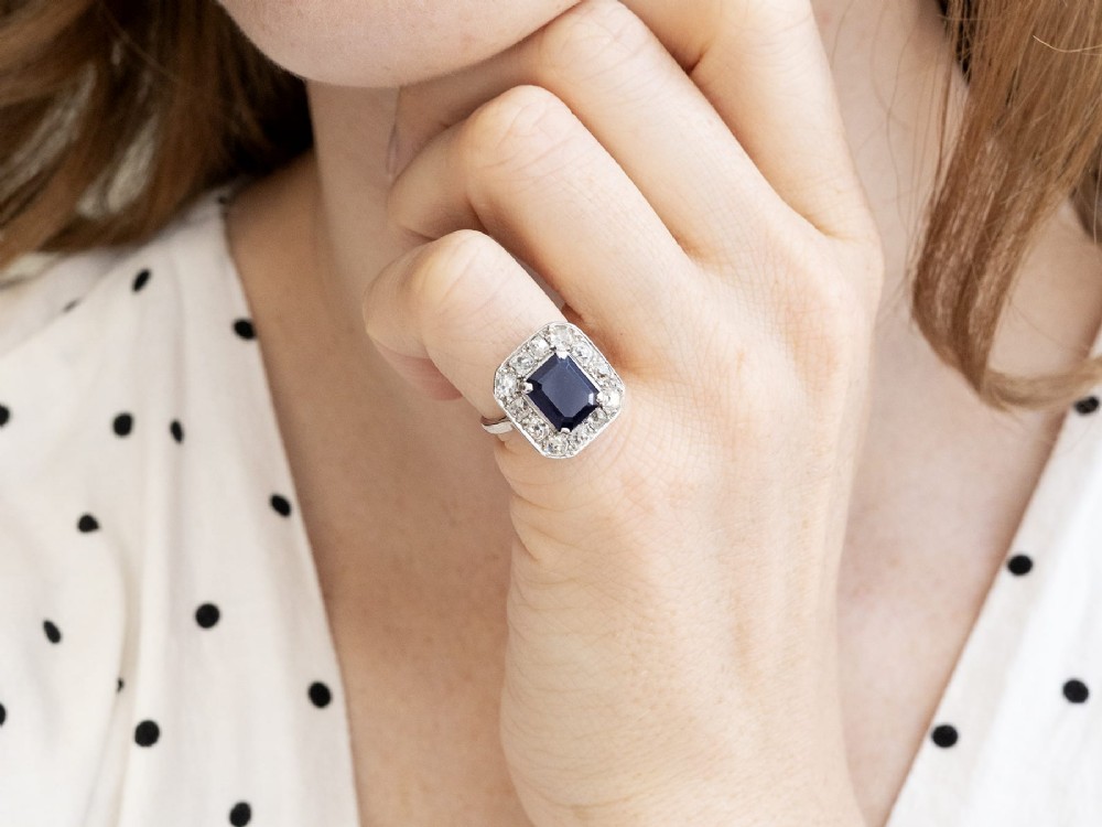 An Art Deco platinum cluster ring featuring a central rectangular blue sapphire surrounded by twelve old-cut diamonds with millegrain edges. View V