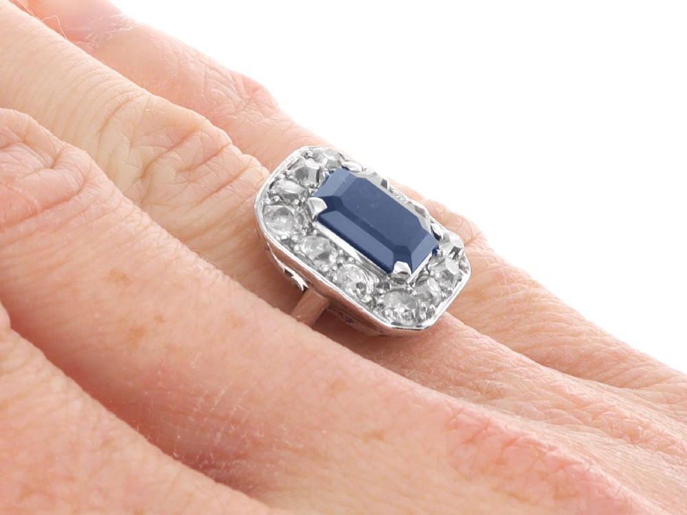 An Art Deco platinum cluster ring featuring a central rectangular blue sapphire surrounded by twelve old-cut diamonds with millegrain edges. View XI