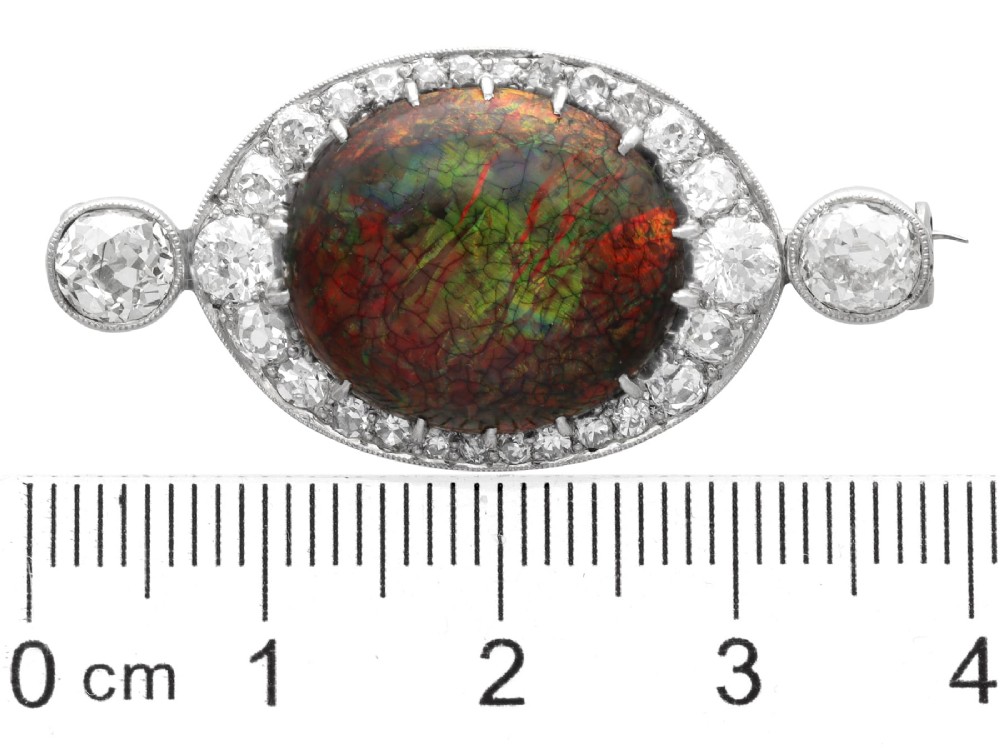 An Edwardian platinum brooch featuring an oval cabochon boulder opal surrounded by a halo of diamonds and two outer accent diamonds. View IX