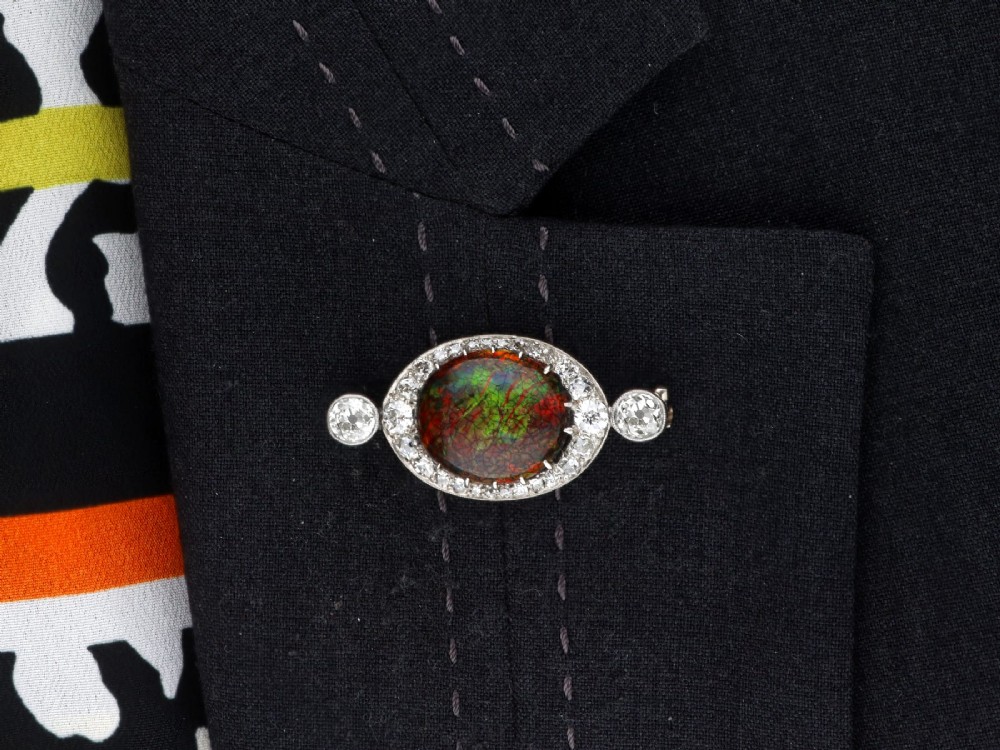 An Edwardian platinum brooch featuring an oval cabochon boulder opal surrounded by a halo of diamonds and two outer accent diamonds. View X