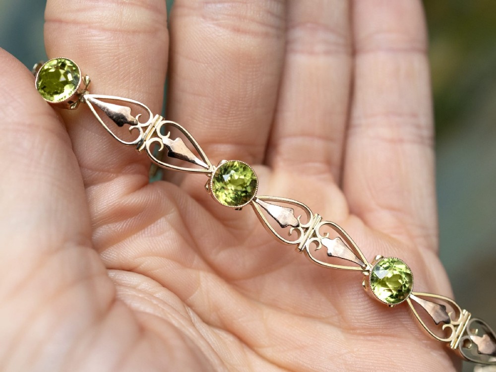 An antique Art Nouveau 9ct yellow gold bracelet featuring round-cut green peridots held in collet settings between openwork scroll links. View V