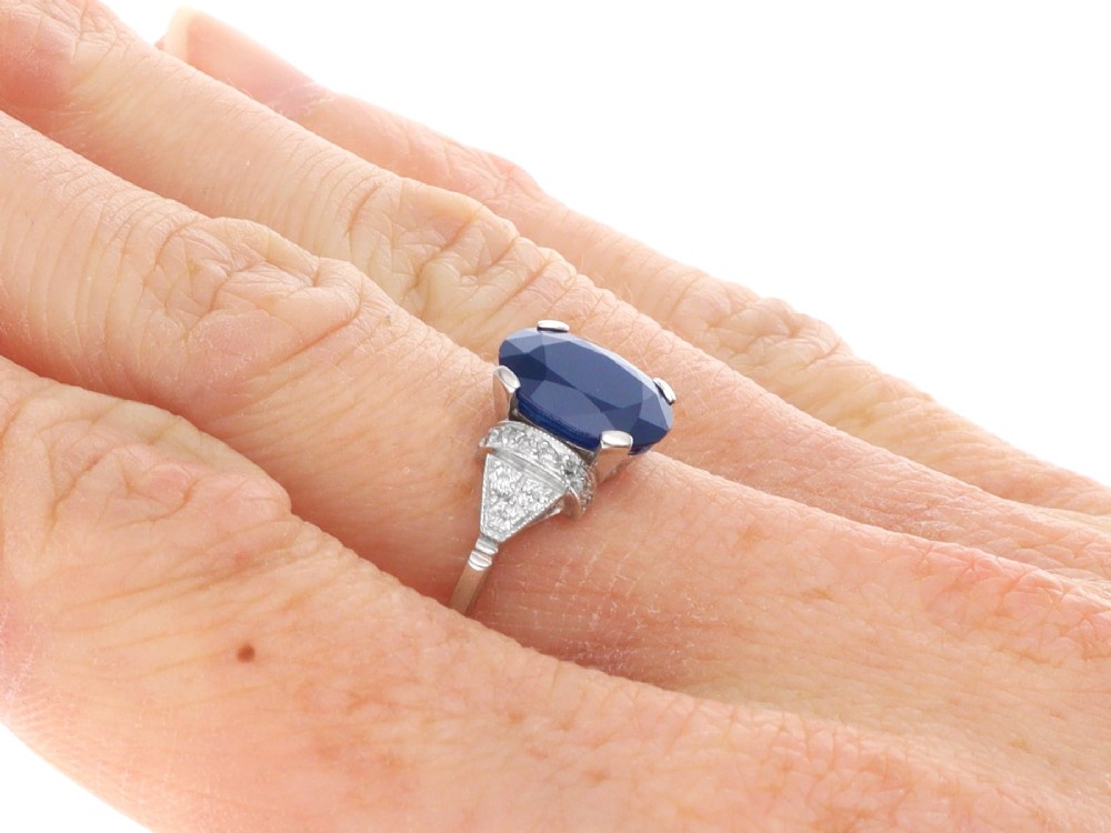An Art Deco platinum ring featuring a centrally set oval blue sapphire flanked by diamond-set geometric shoulders. View IX