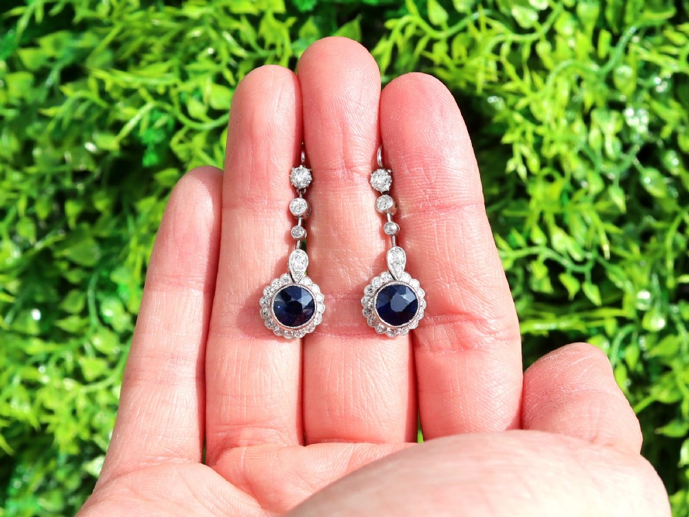 A pair of antique Art Deco platinum drop earrings featuring round dark blue sapphires encircled by diamond halos suspended from articulated diamond links. View II