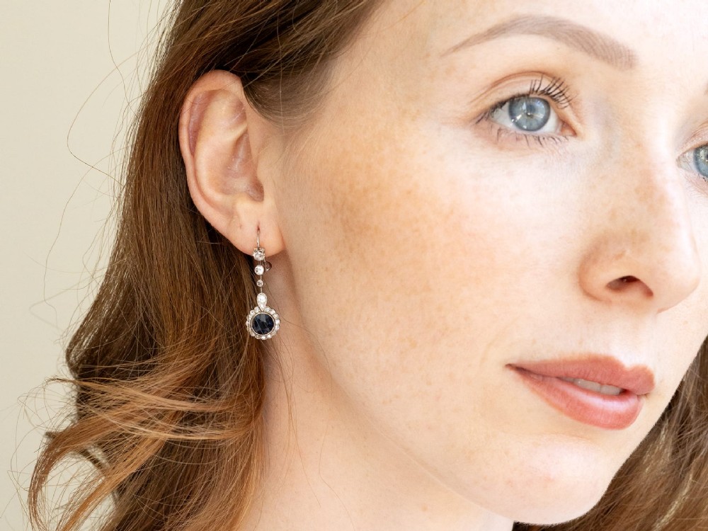 A pair of antique Art Deco platinum drop earrings featuring round dark blue sapphires encircled by diamond halos suspended from articulated diamond links. View X