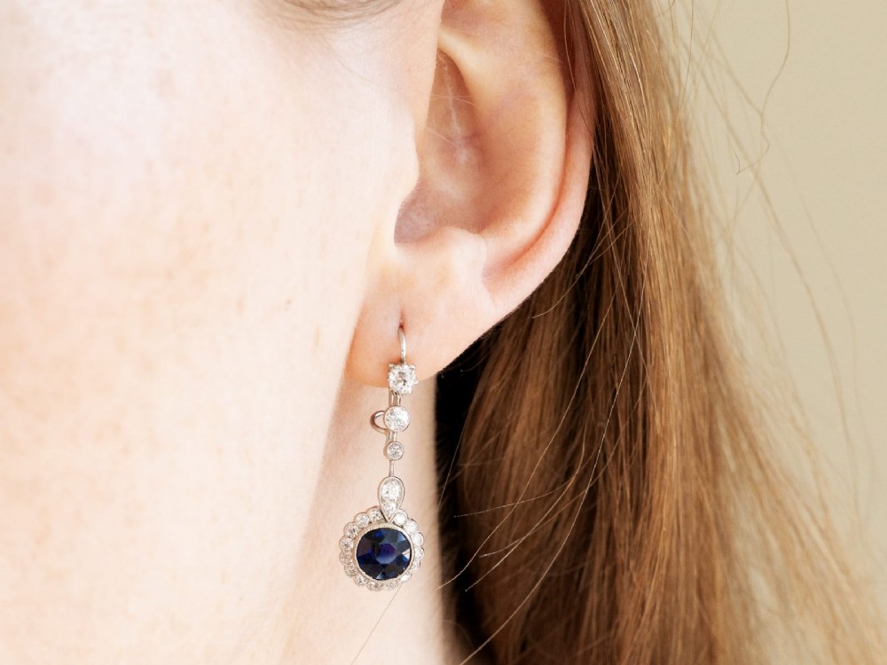 A pair of antique Art Deco platinum drop earrings featuring round dark blue sapphires encircled by diamond halos suspended from articulated diamond links. View XI