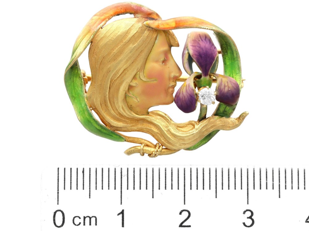 An Art Nouveau 18ct yellow gold brooch detailing a maiden profile framed by green enamel foliage and a purple enamelled iris set with a single diamond. View X
